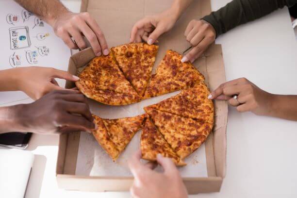 Many people grabbing their own slice of pizza from a cardboard pizza box. The pizza grease contaminates the cardboard for recycling purposes