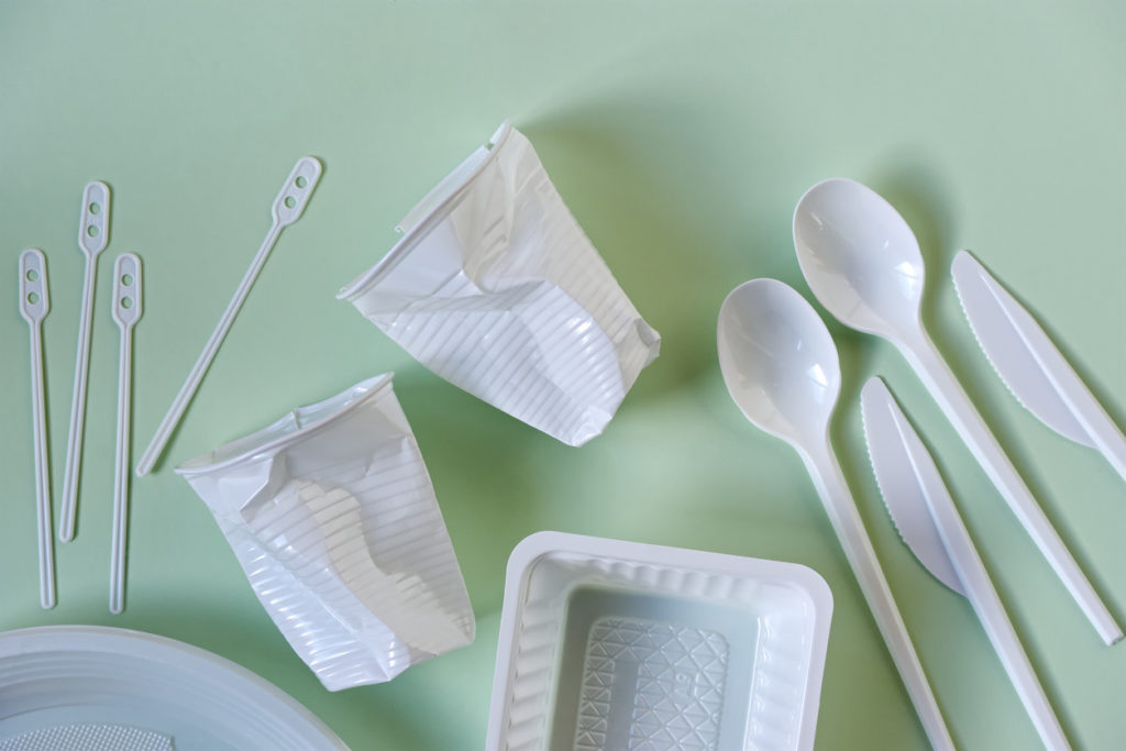 Two smashed white plastic coffee cups, spoons, knives, stirrers, plate and box on a light green background. Zero waste, plastic free, stop pollution, ecological concept.