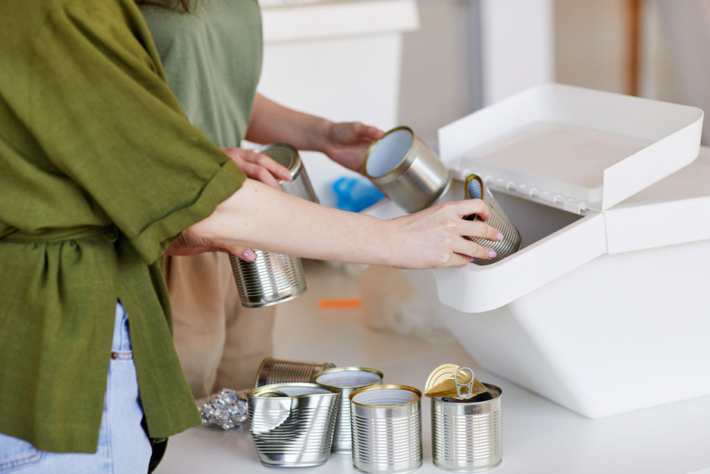 Side view close up of unrecognizable women putting discarded metal cans in plastic container while sorting waste materials for recycling, copy space